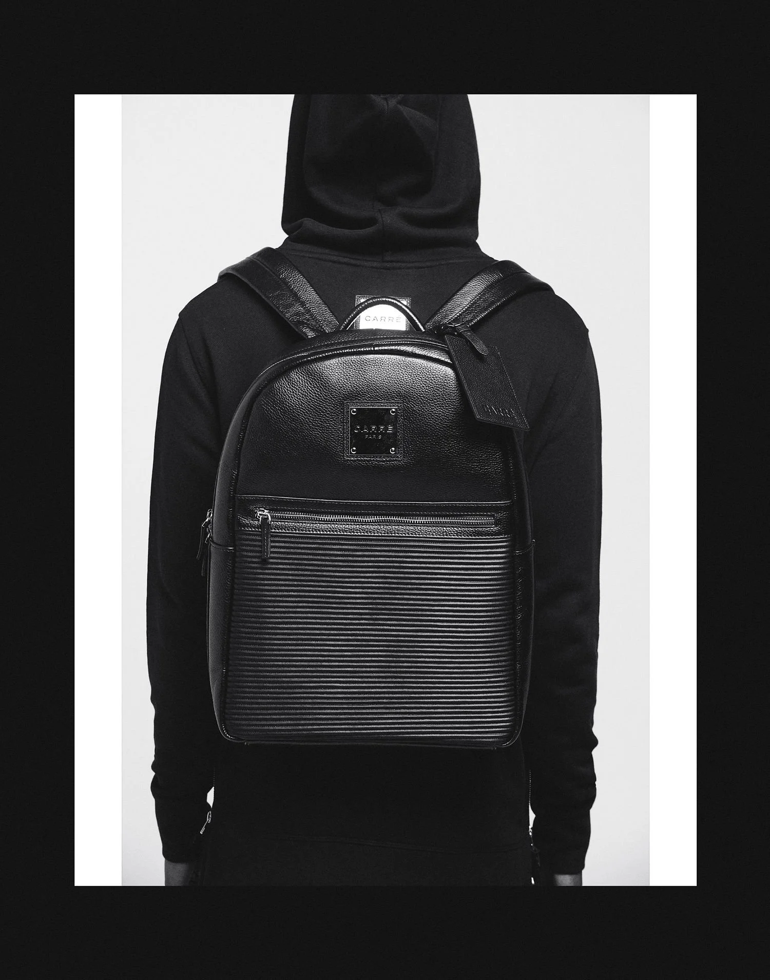 Black backpack with ribbed front pocket on a person dressed in black with a black hoodie, against a gray and black background.