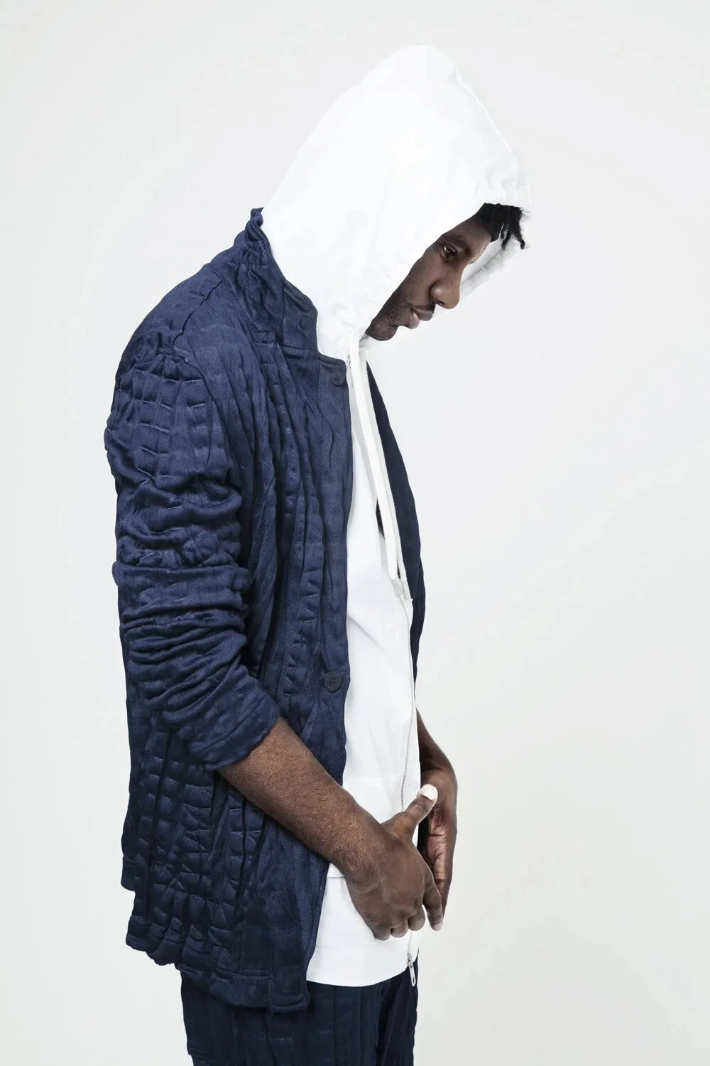 A man wearing a white hoodie and a blue quilted jacket, looking down with hands in pockets, against a plain white background.