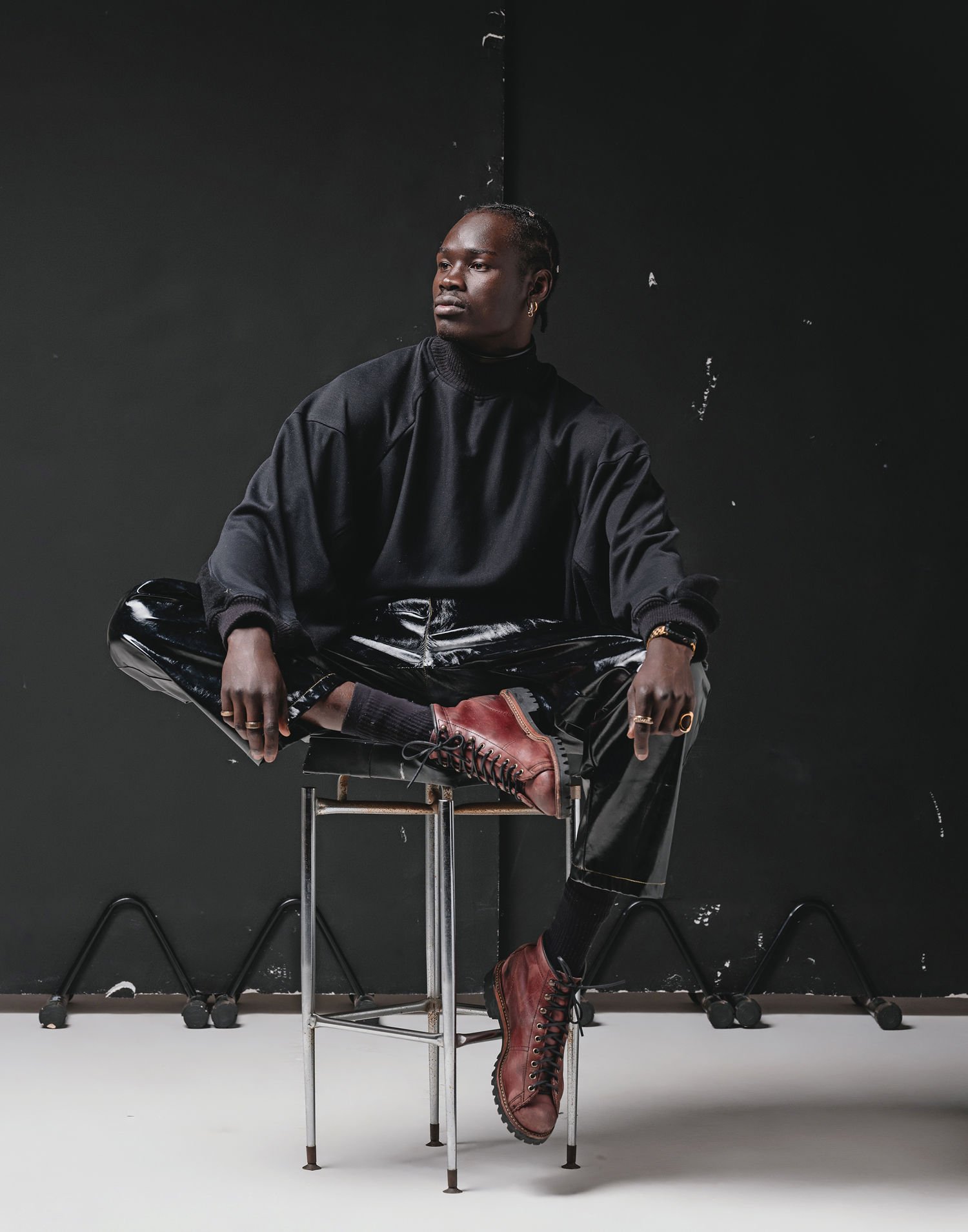 A person with dark skin sitting cross-legged on a chair in front of a black wall, wearing a black sweatshirt, shiny black pants, and red lace-up boots.