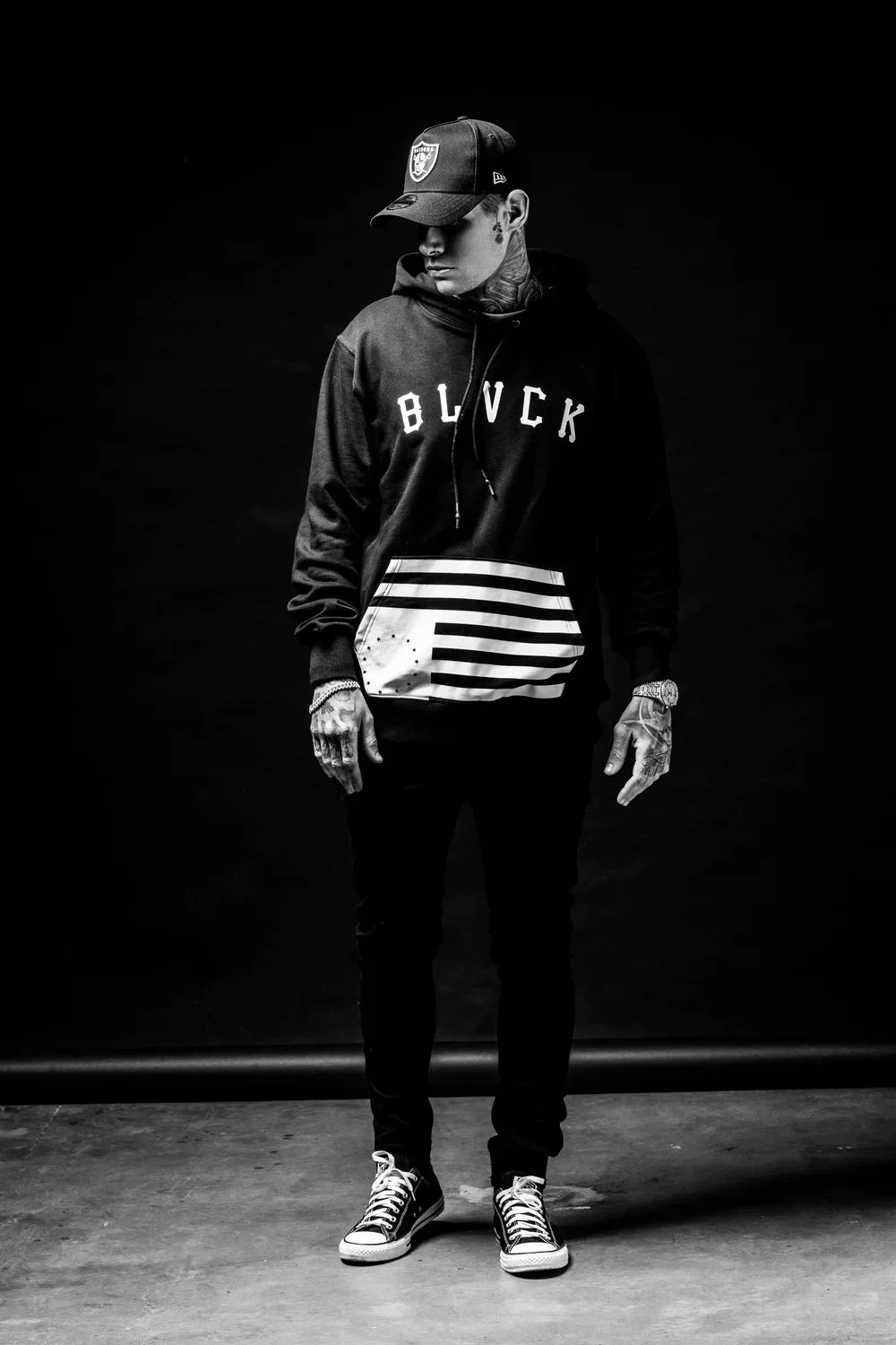 A young man with tattoos and earrings wearing a cap, hoodie with the word "BLACK" on it, striped pouch, and sneakers, standing against a dark background.