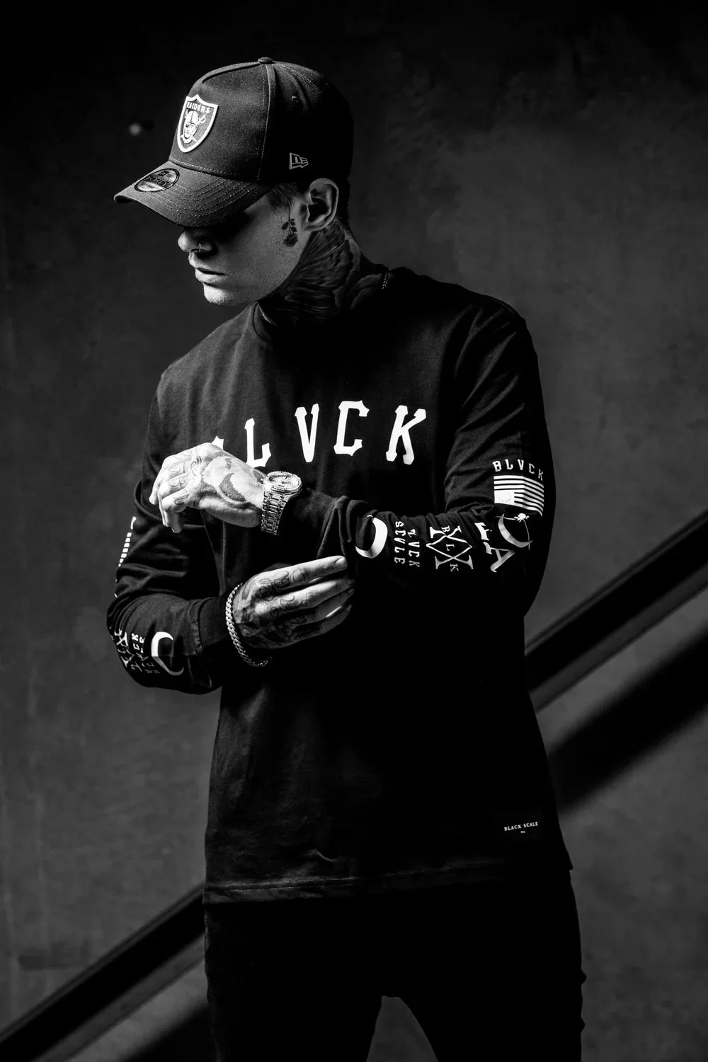 A man wearing a baseball cap, tattoos, and a long sleeve black shirt with various text and patches, standing against a dark background.