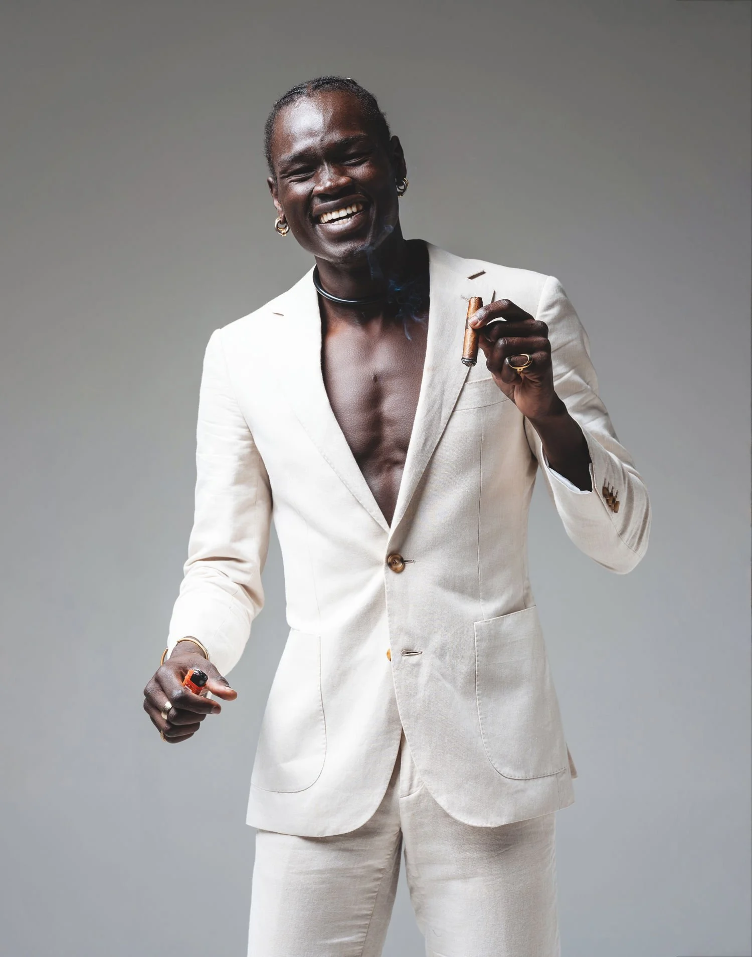 A man with dark skin, dressed in a cream-colored suit with an open shirt, smiling and holding a cigar and a lighter, standing against a plain gray background.