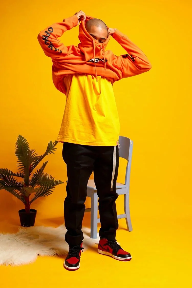 Person in a yellow and orange hoodie with a bald head, standing in front of a yellow background, with a blue chair, potted plant, and white fur rug nearby.