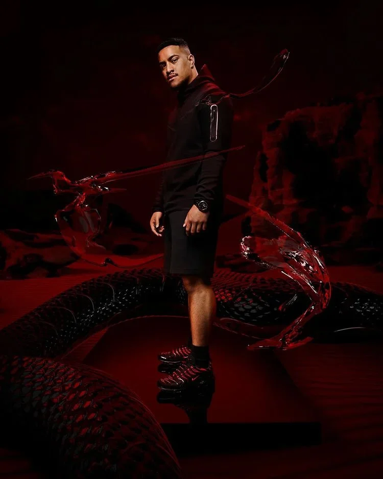 A man wearing a black jacket, shorts, and a watch standing on a black platform with a red and black abstract background, featuring a large serpent and swirling design elements.