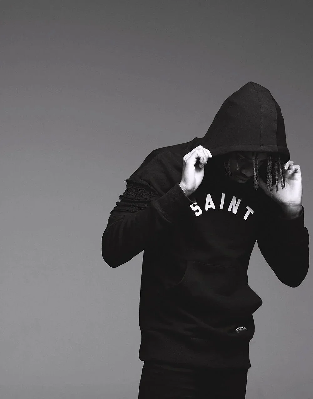 A person wearing a black hoodie with the hood up, partially covering their face, and a black sweatshirt with 'SAINT' written across the chest. The person is holding the hoodie with both hands, standing against a plain grey background.