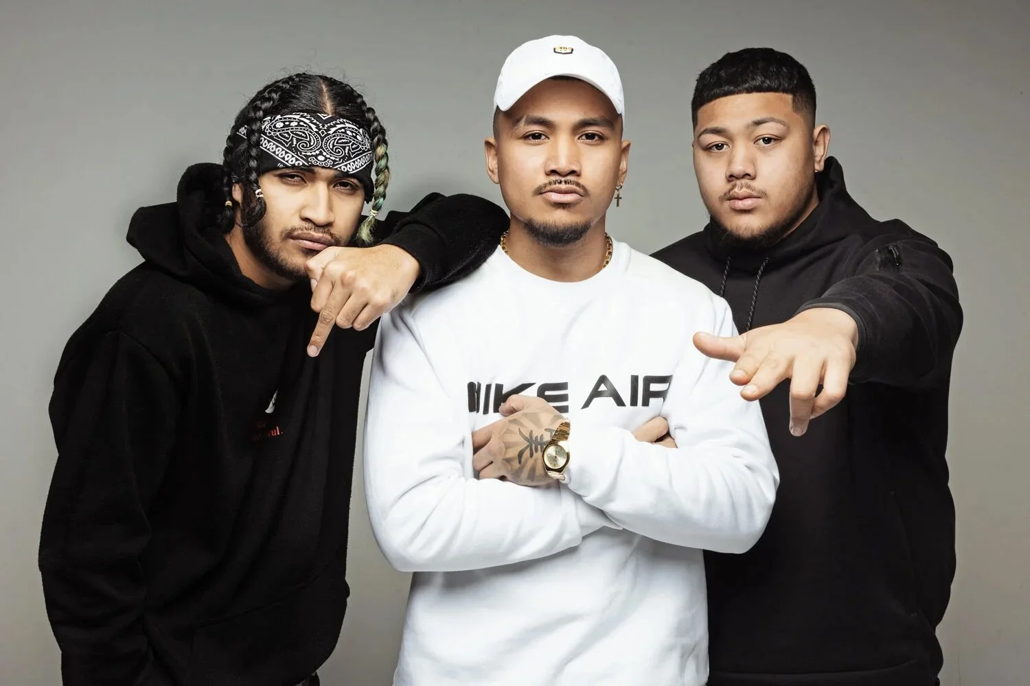 Three young men standing close together against a plain background, wearing hoodies and casual clothing, with confident expressions.