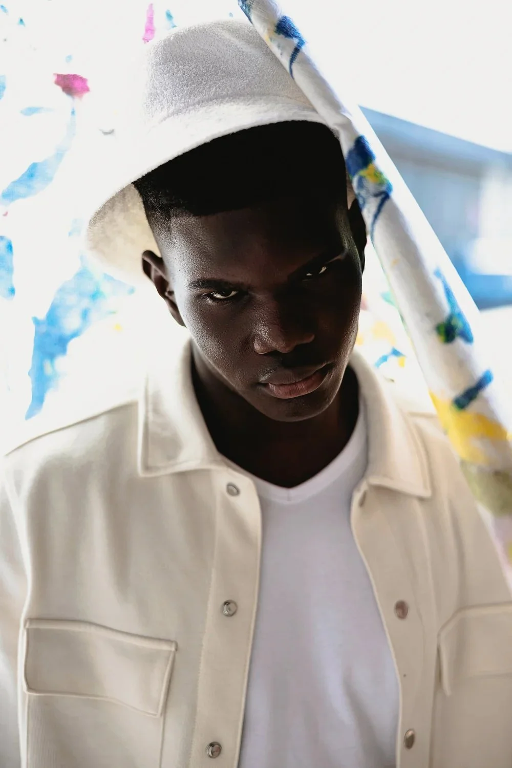 A young Black man with dark skin wearing a white shirt and a fedora hat, holding a colorful umbrella with a white background.