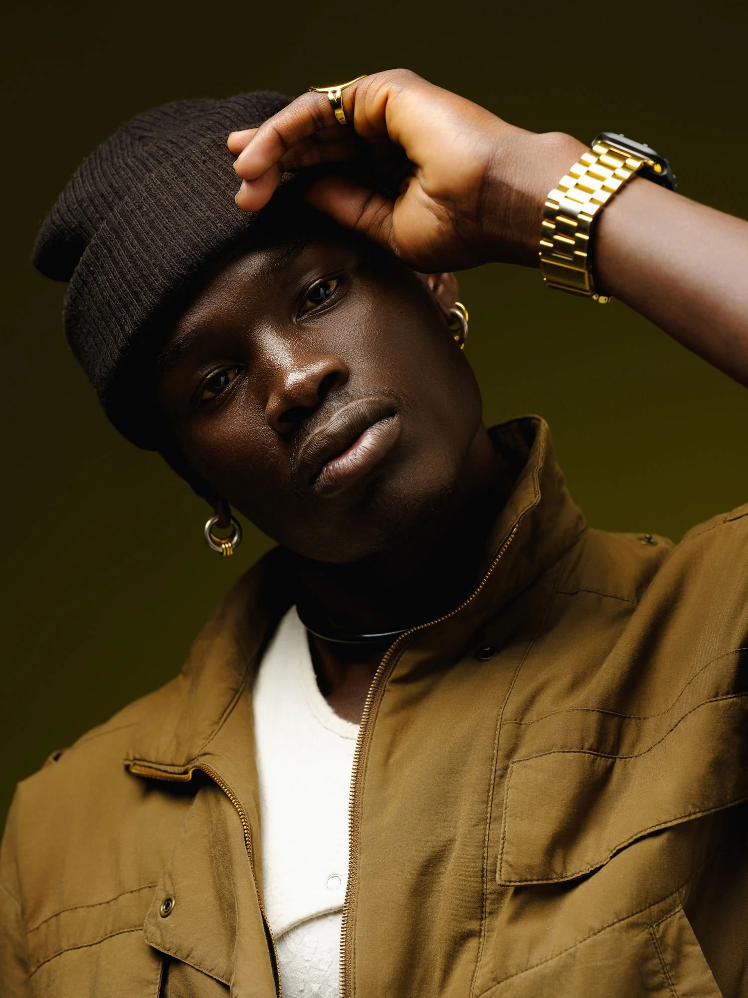 A young African American person wearing a black beanie, brown jacket, white shirt, gold jewelry, and a wristwatch, posing with their hand on their head against a dark green background.