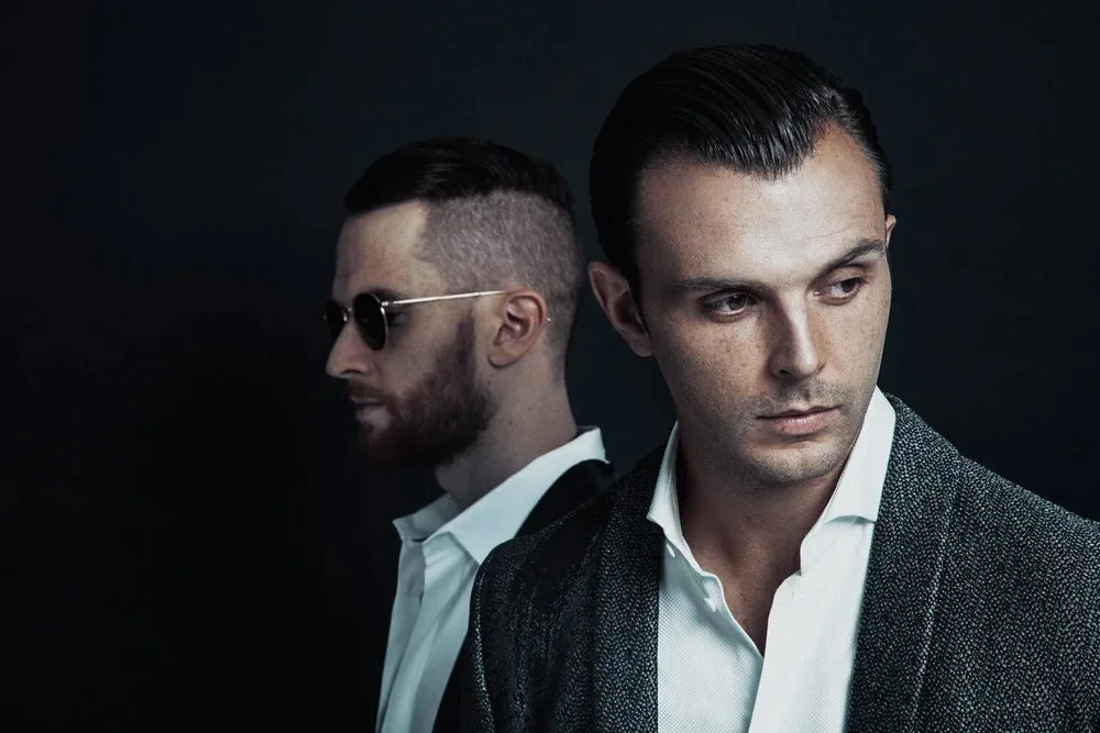 Two men in stylish clothing against a dark background, one with sunglasses looking away, the other looking to the side.