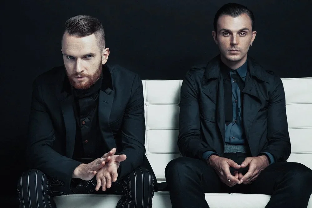 Two men sitting on a white modern bench against a black background, both dressed in dark, fashionable clothing, with serious expressions.