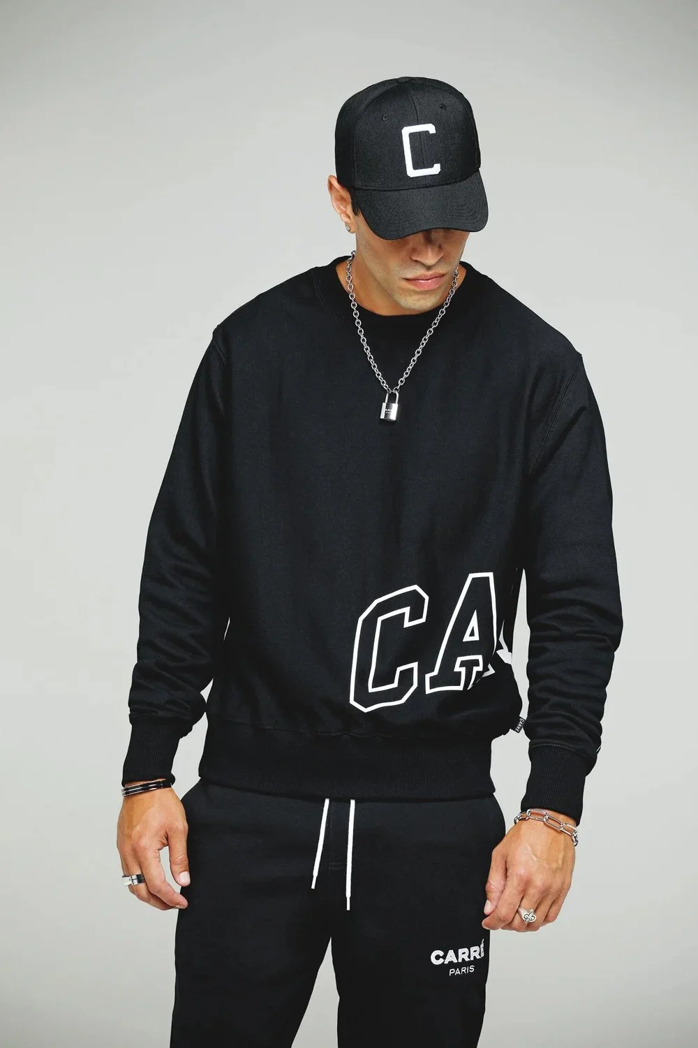 A man dressed in black athletic wear, including a black cap with a white 'C', a black sweatshirt with 'C A' lettering, and black pants with 'CARRI PARIS' writing, stands against a plain background.