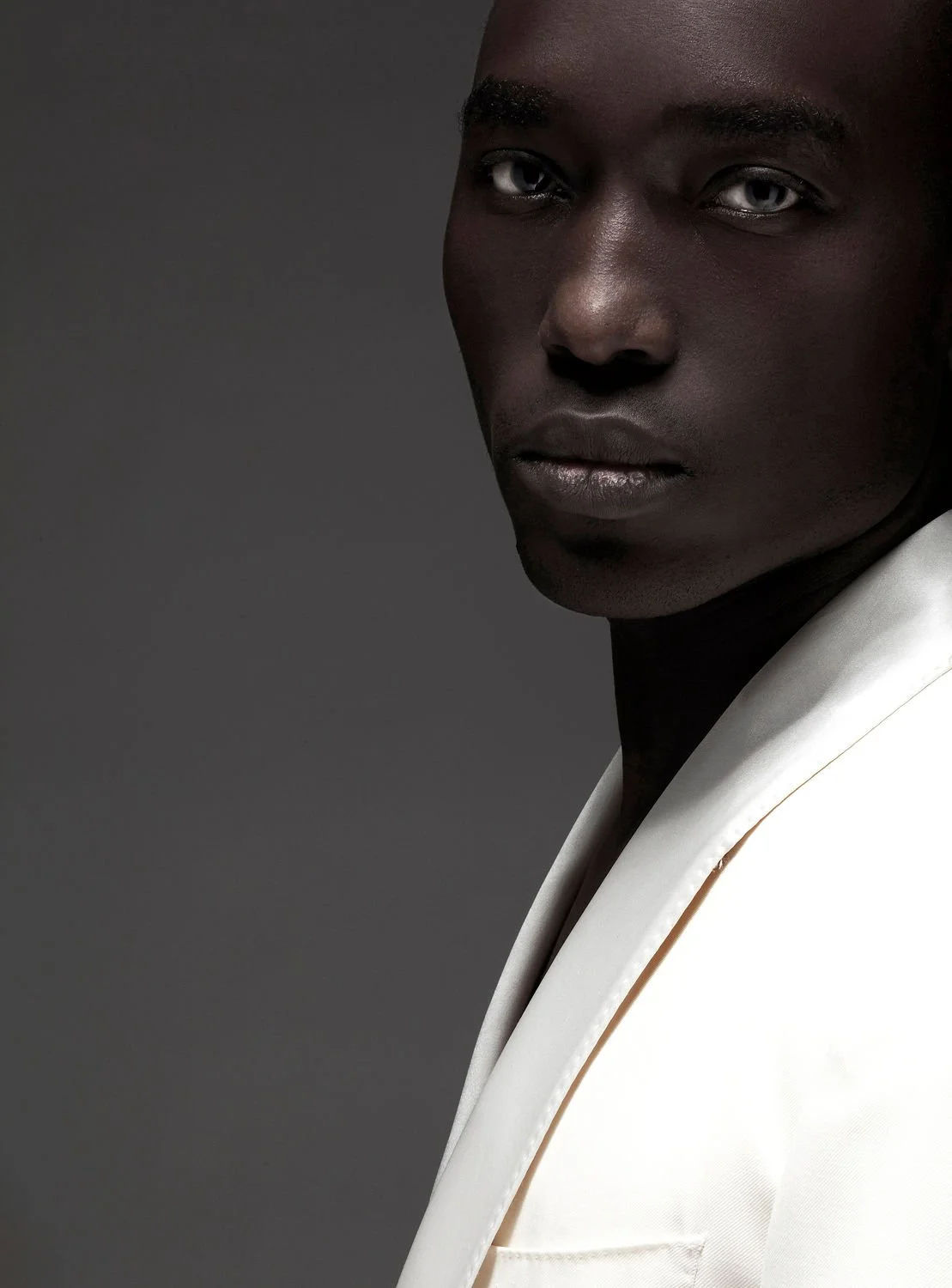 Close-up of a dark-skinned man with silver eyes, wearing a white suit, facing slightly to the right against a dark background.