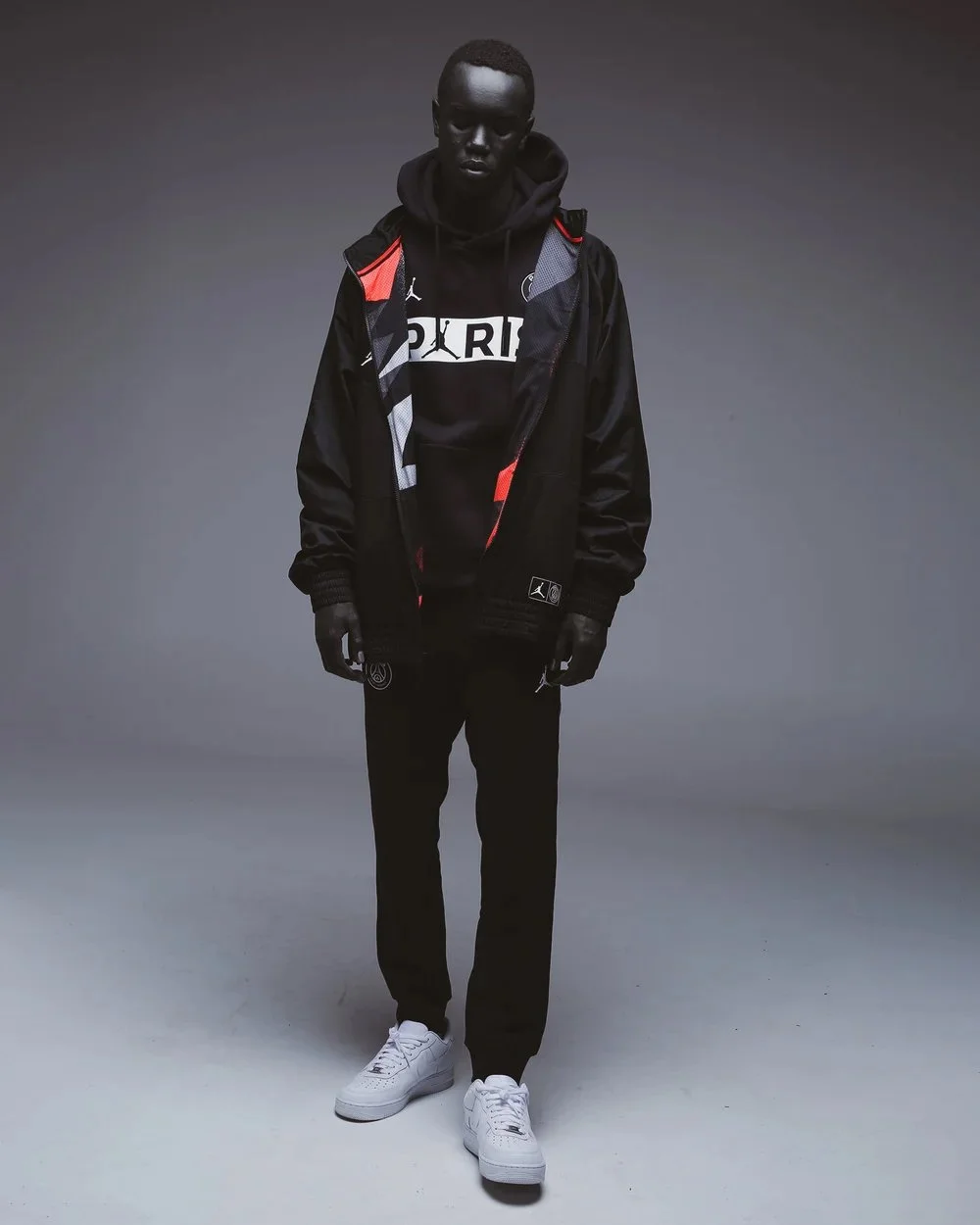 A young man with dark skin standing against a plain gray background. He is wearing a black hoodie, a black and red jacket with a hood, black pants, and white sneakers. He has a black backpack on his shoulders and is looking down with his hands relaxe