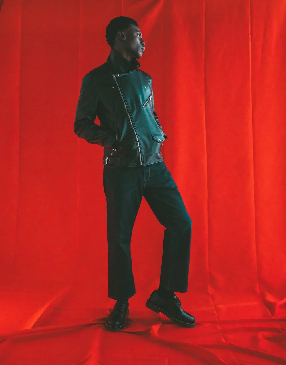 A young man with dark skin and short curly hair standing in front of a bright red textured background. He is wearing a black leather jacket, black pants, and black boots. His hands are in his jacket pockets, and he is looking to his left.