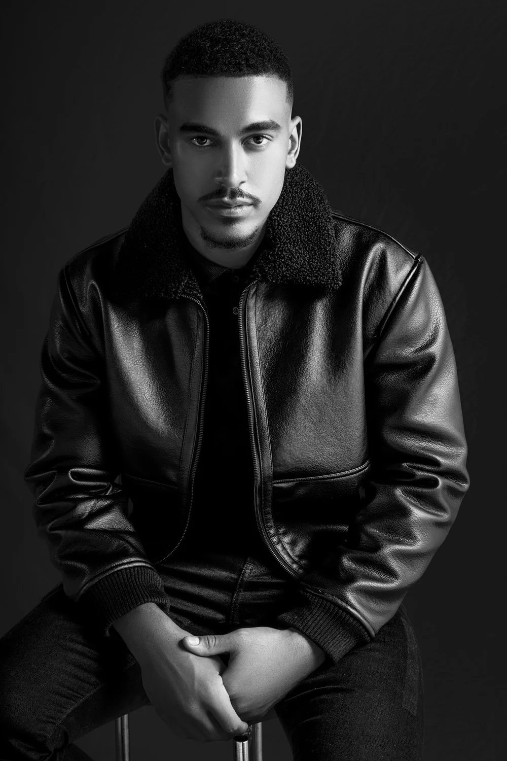 Black and white portrait of a man wearing a leather jacket with a shear collar, seated against a dark background.