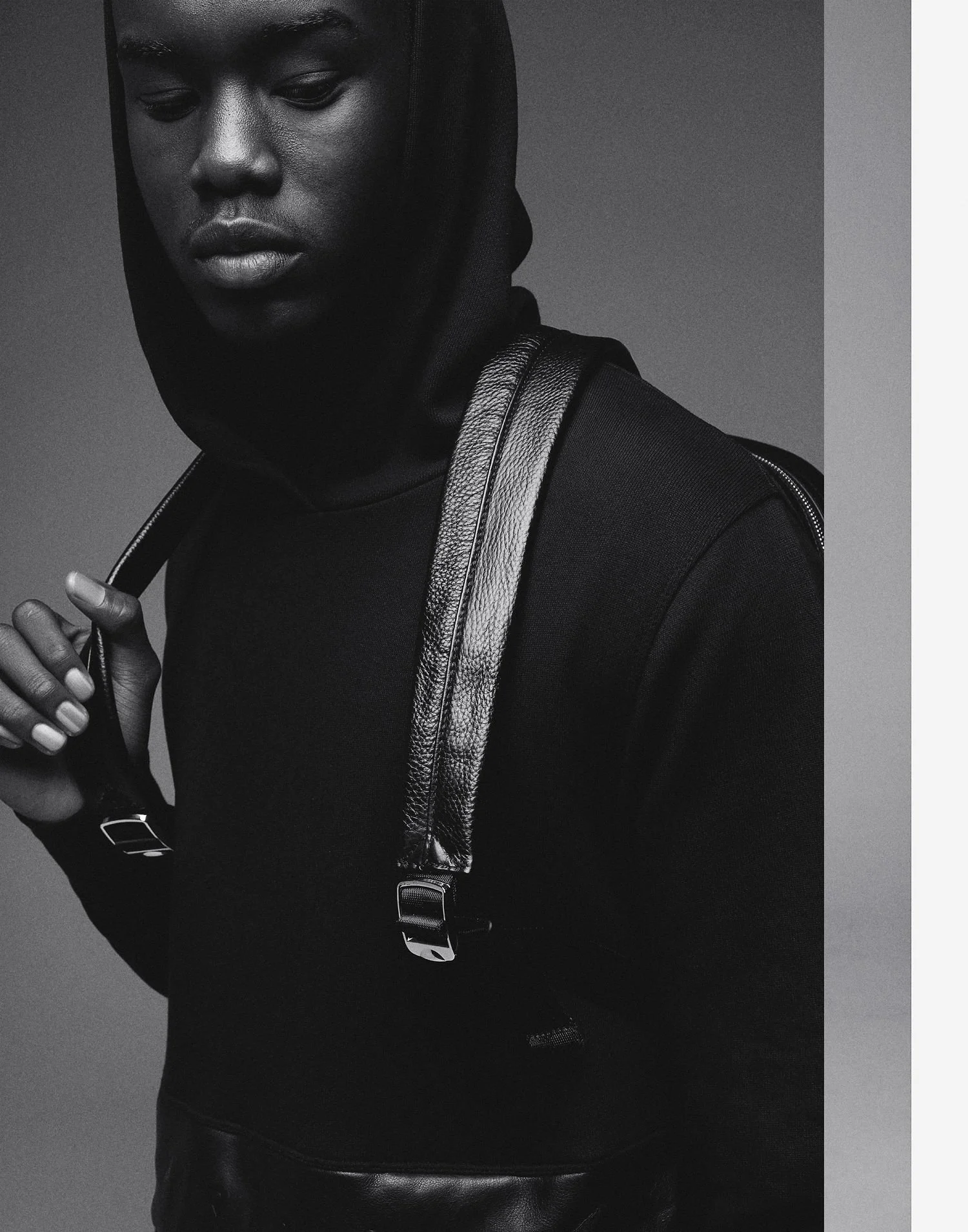 A black and white photo of a man in a black hoodie with a backpack, looking down and holding the strap of the backpack.