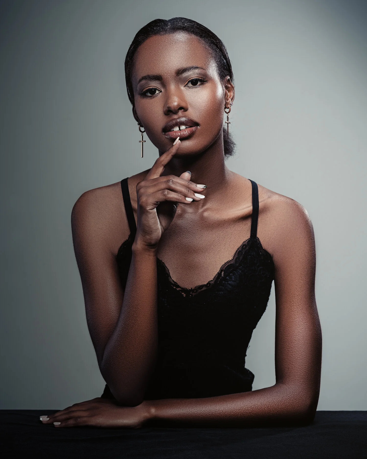A woman with dark skin and short hair wearing a black lace camisole and cross earrings, posing with her hand near her face against a gray background.