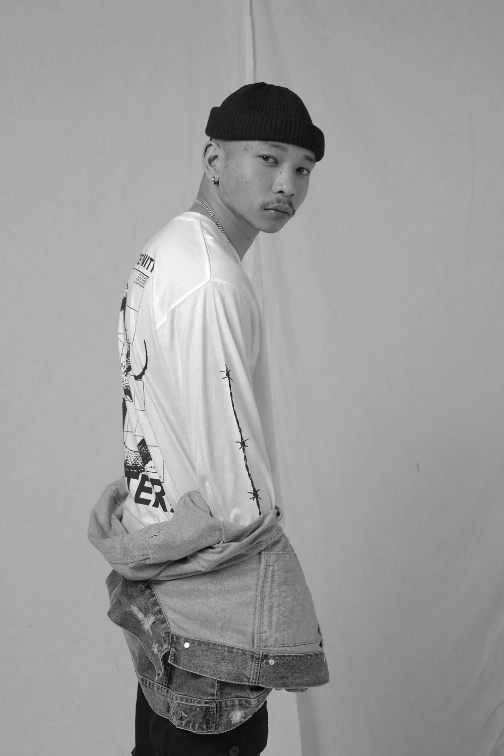 Black and white photo of a young man wearing a black beanie, a patterned long-sleeve shirt, and ripped jeans, standing against a plain wall, looking at the camera with a serious expression.