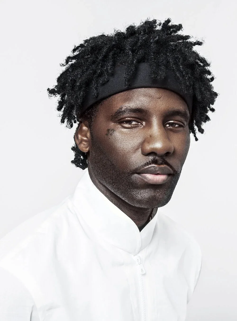 A man with dark skin, curly black hair, and a black headband, wearing a white shirt with a high collar, indoors against a plain white background.