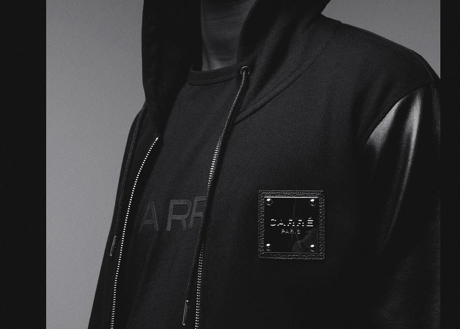 Close-up of a person wearing a black jacket with leather sleeves and a black shirt with a logo, against a gray background.