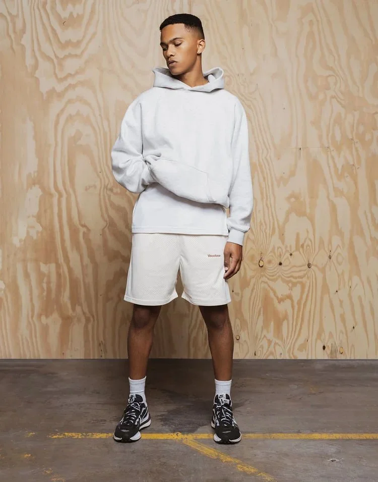Young man wearing a light gray hoodie, white sports shorts, white socks, and black sneakers standing against a wooden panel background in an indoor setting.