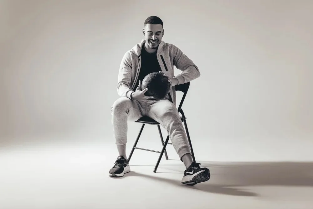 A young man with a smile, sitting on a chair, holding a basketball, wearing casual athletic clothing in a studio with a neutral background.