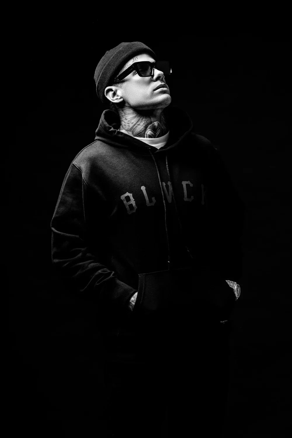 Black and white photo of a man wearing sunglasses, a beanie, and a hoodie, with his hands in the pockets, looking upwards against a dark background.