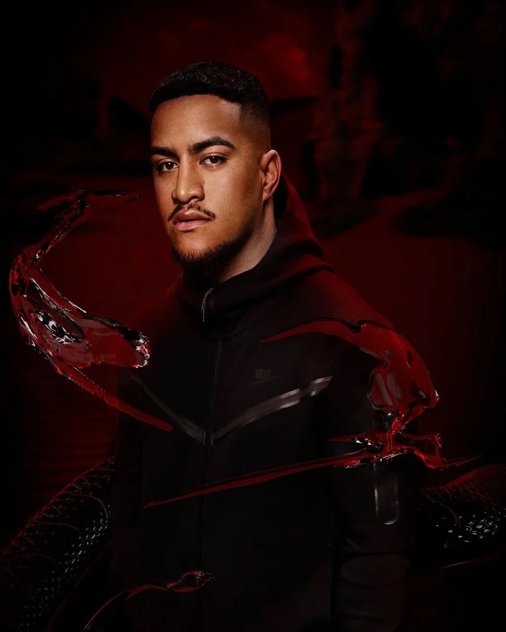 Portrait of a young man with short dark hair and a goatee, wearing a black zip-up hoodie, with a dark red background and red and black digital effects.