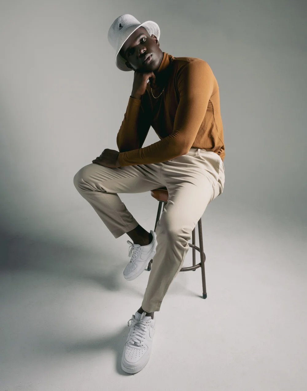 A man sitting on a stool, wearing a white hat, brown long sleeve shirt, cream pants, and white sneakers, posing with one hand resting on his chin against a plain, light gray background.