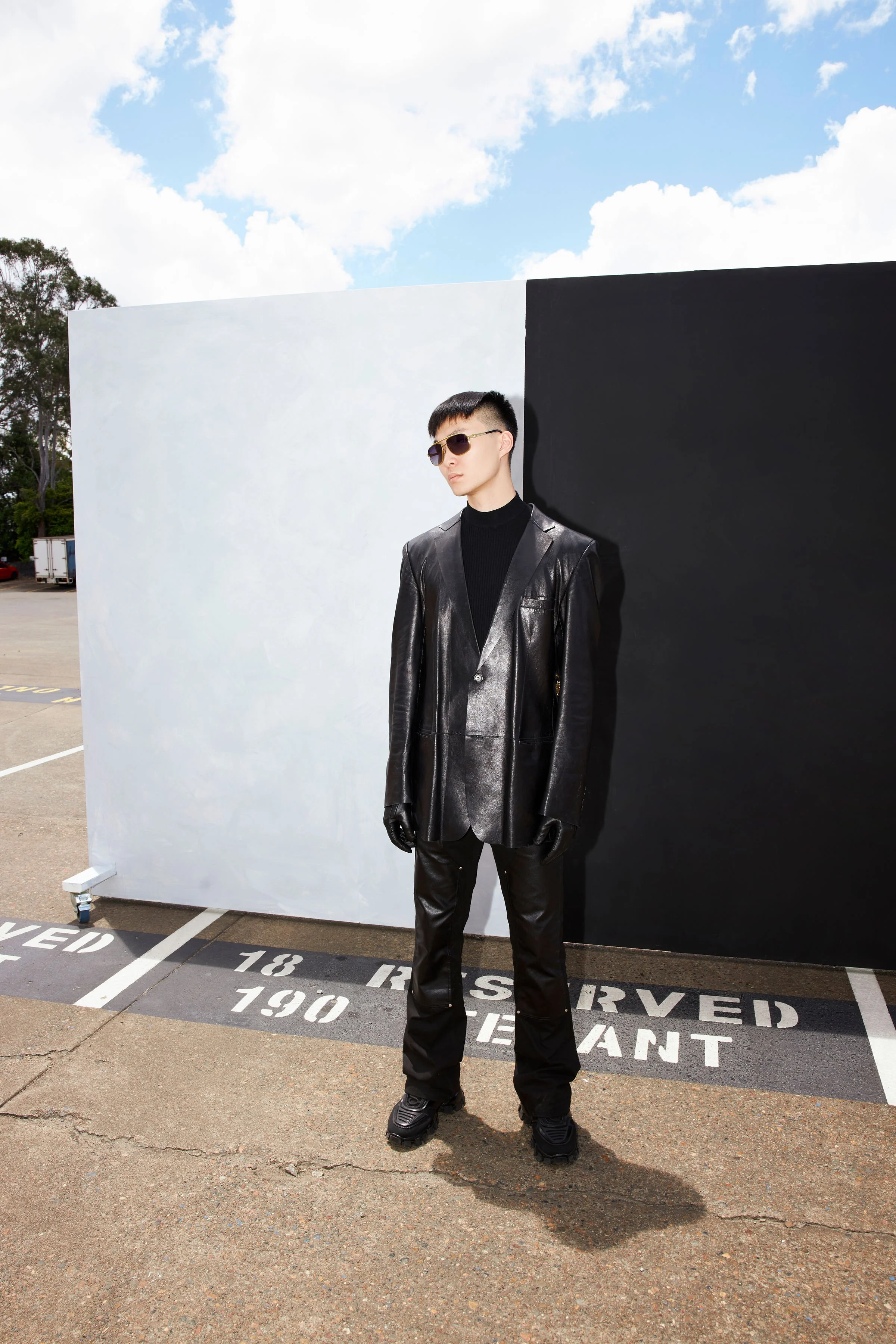 Young man in black leather suit and sunglasses standing outdoors in front of black and white partition wall under partly cloudy sky.