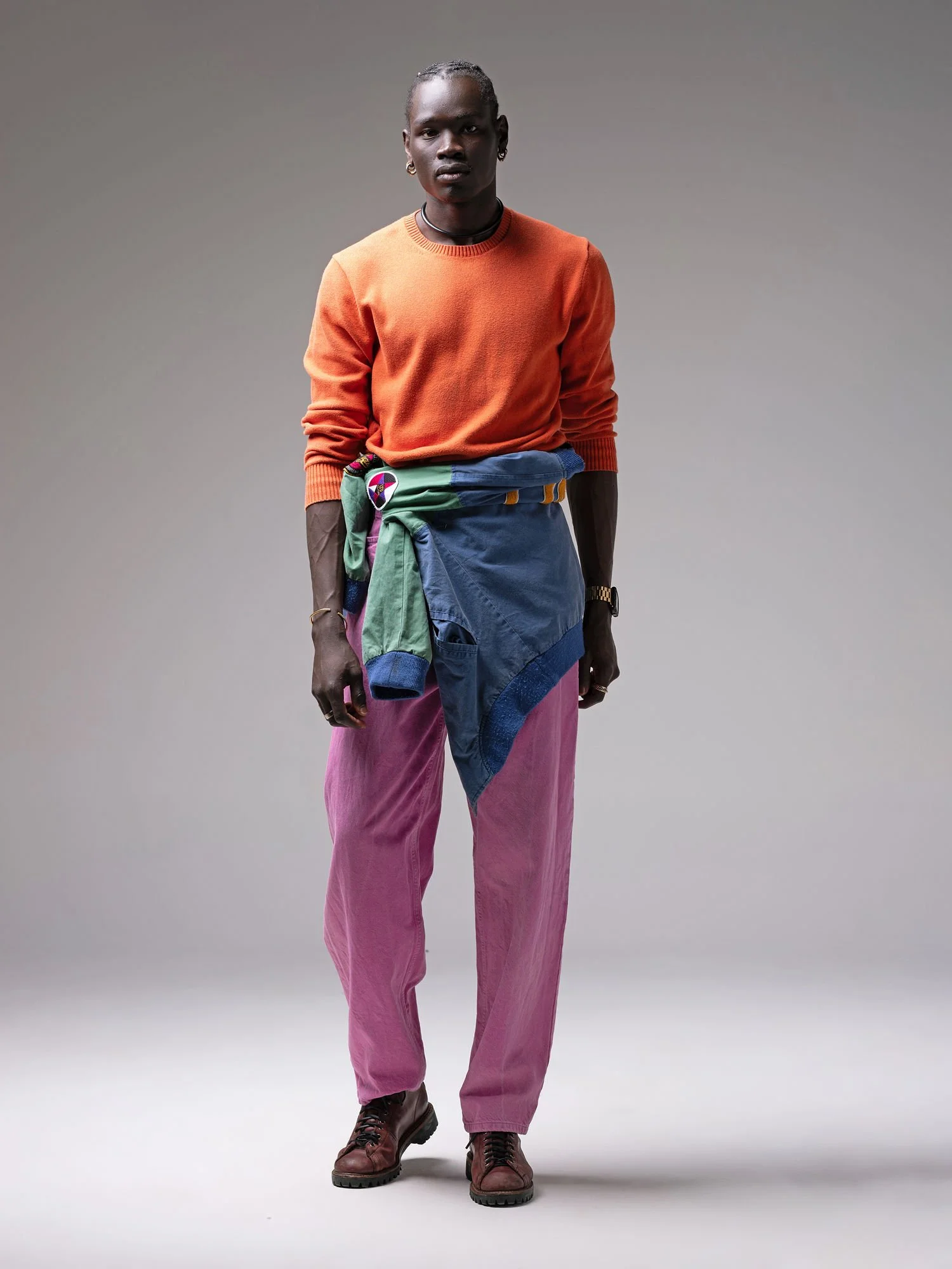A person standing against a plain gray background, wearing a burnt orange sweater, pink pants, brown boots, and a multicolored jacket tied around the waist.