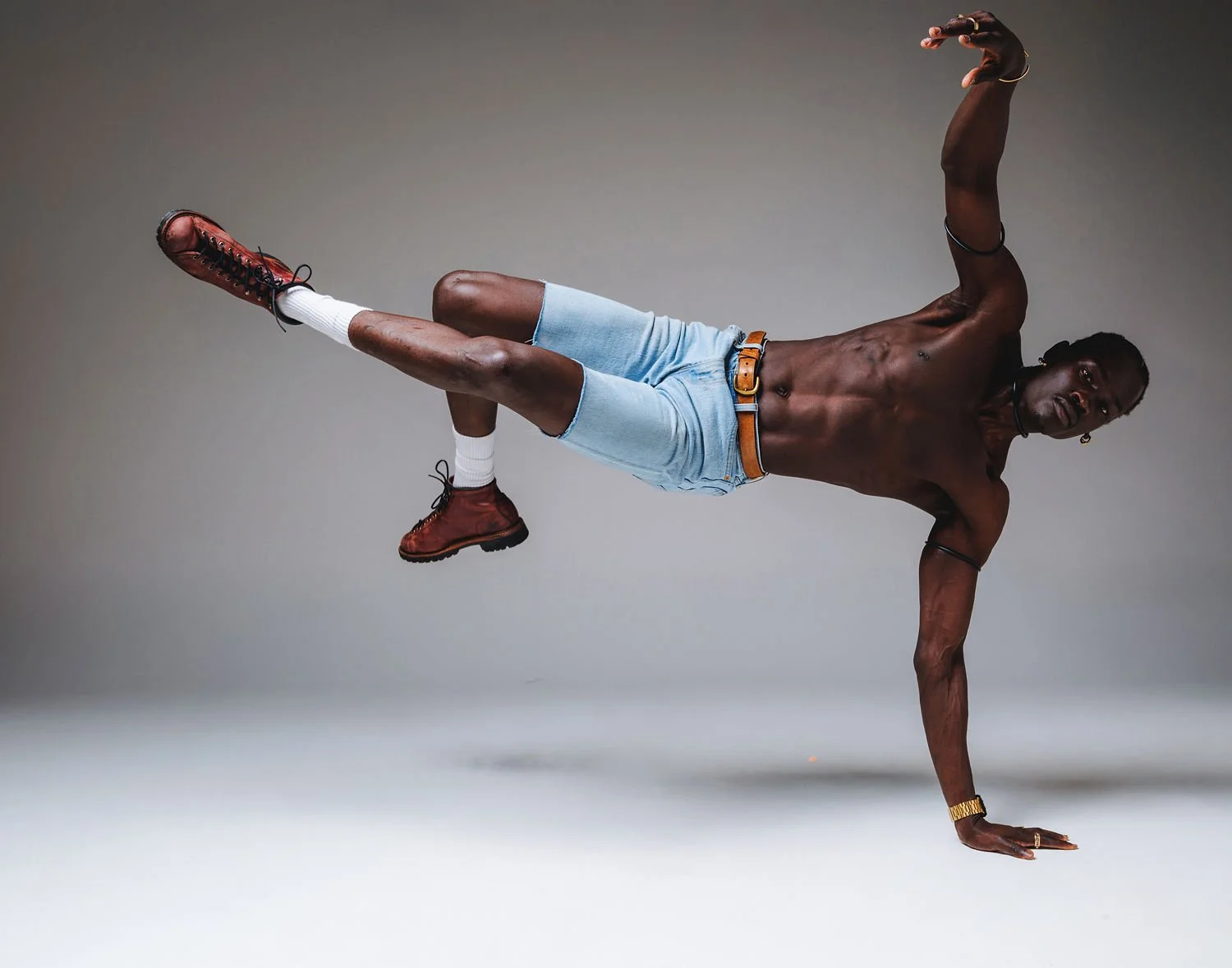A shirtless man wearing denim shorts, red boots, white socks, and gold jewelry performs a one-handed balancing pose against a plain background.