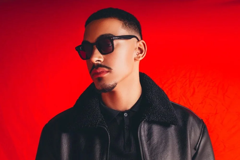 A stylish man wearing sunglasses and a black leather jacket with a fur collar, posing against a red background.