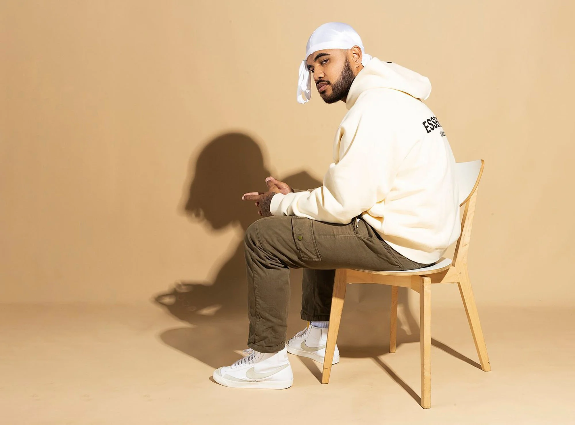 A man sitting on a wooden chair against a beige background, wearing a white hoodie with black text on the back, brown pants, white sneakers, and a white durag, looking at the camera with a serious expression.