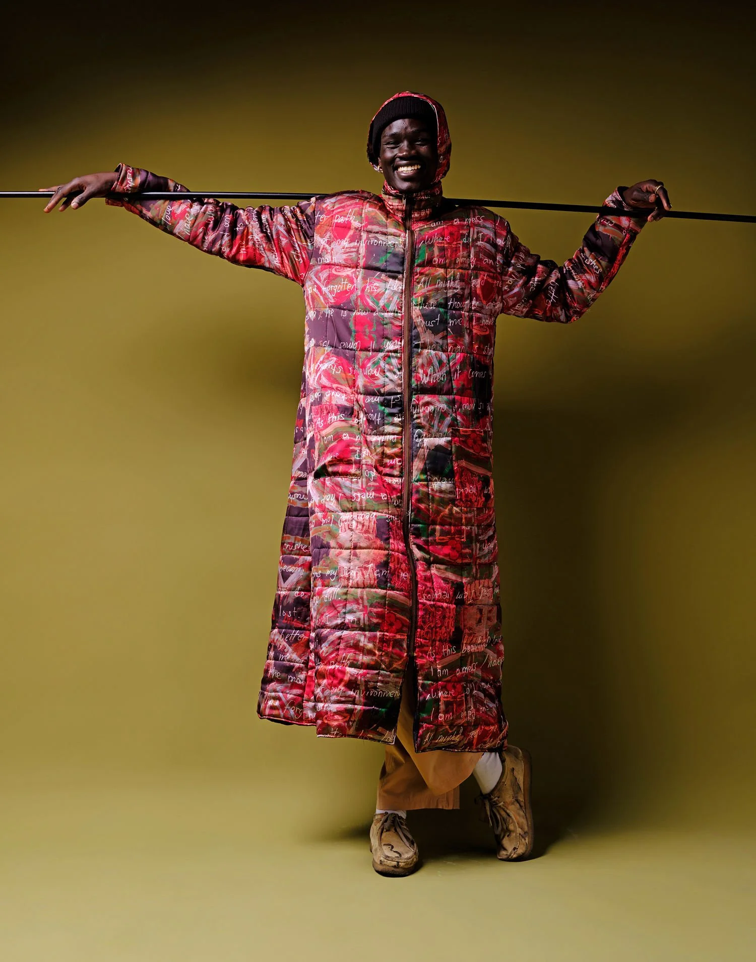 Person in a long, colorful, patterned winter coat, holding a pole behind their neck, smiling, set against a yellow background.