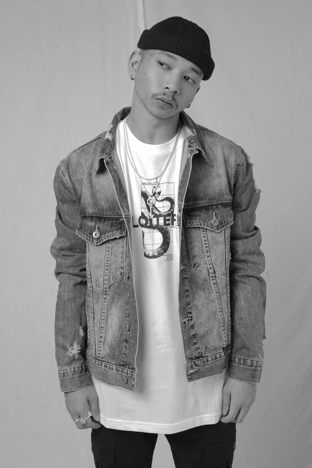 Black and white photo of a young man wearing a beanie, denim jacket, graphic T-shirt, and layered necklaces, posing against a plain wall.