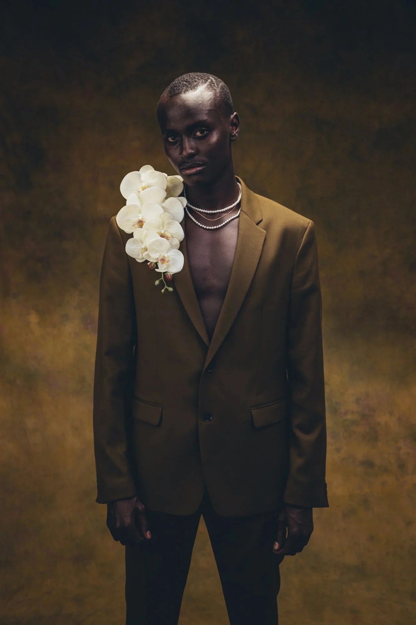 A person with dark skin and short hair wearing a brown suit jacket, layered pearl necklaces, and a large cluster of white orchids on the shoulder, standing against a dark, textured background.