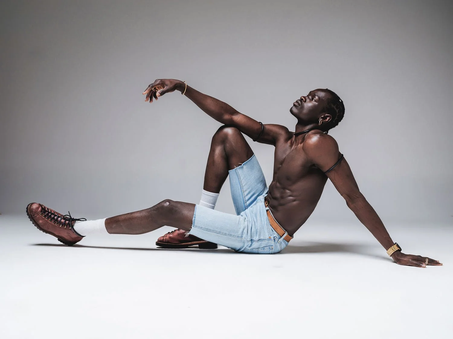 A shirtless man with dark skin, wearing light blue denim shorts, white socks, and brown shoes, sitting on the floor against a plain gray background, with one arm extended and head tilted back.
