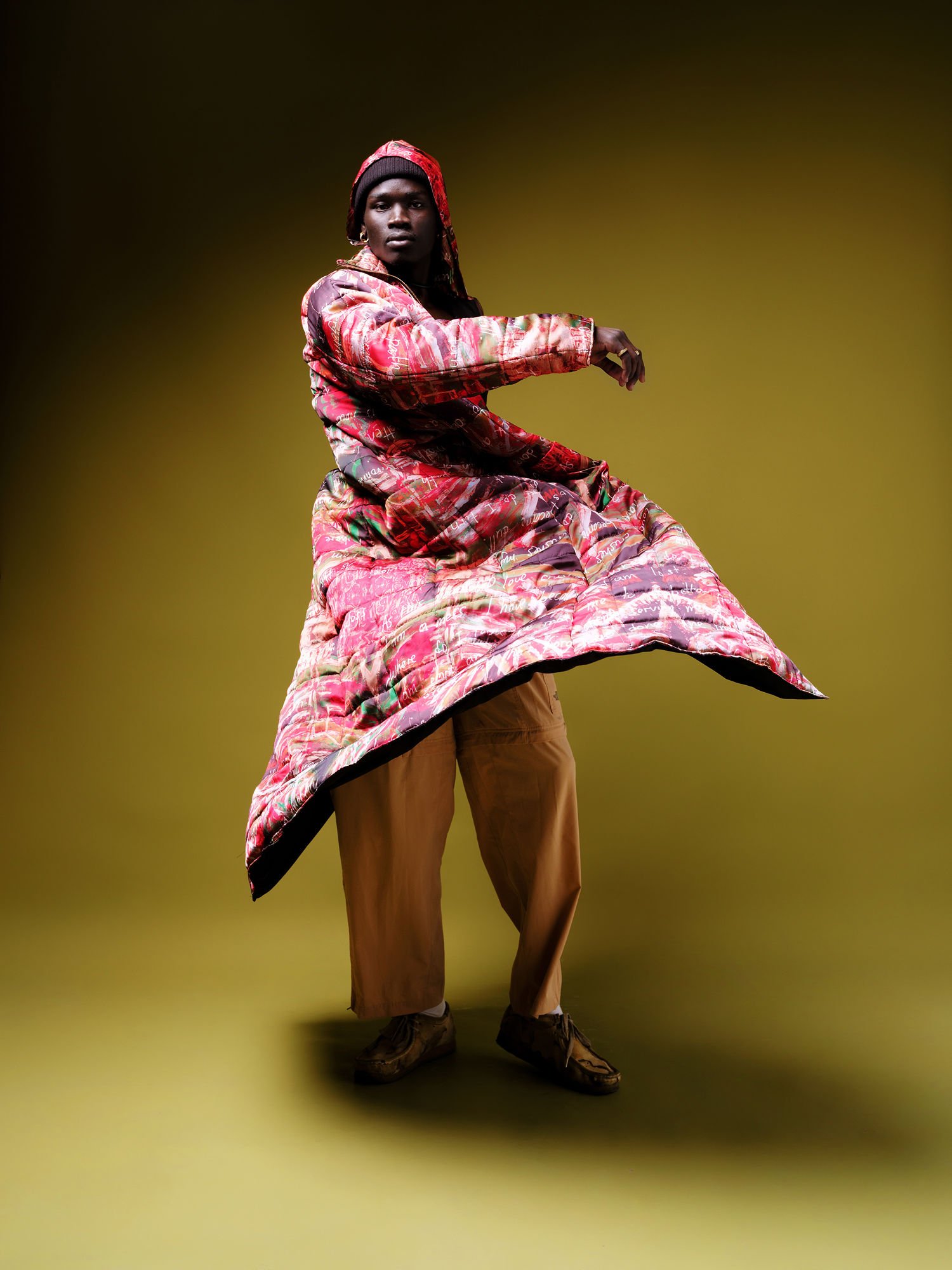 A person wearing a colorful, patterned puffy coat, brown pants, and brown shoes posing against a gradient yellow background.