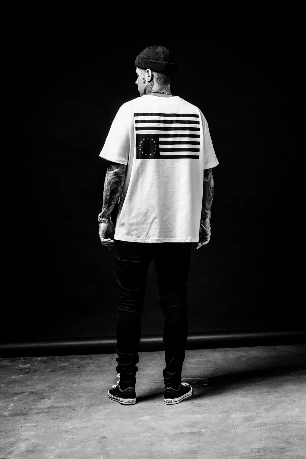 A man with tattoos and a beanie, standing against a dark background, wearing a white t-shirt with a plus sign in a circle and black stripes, and black pants.