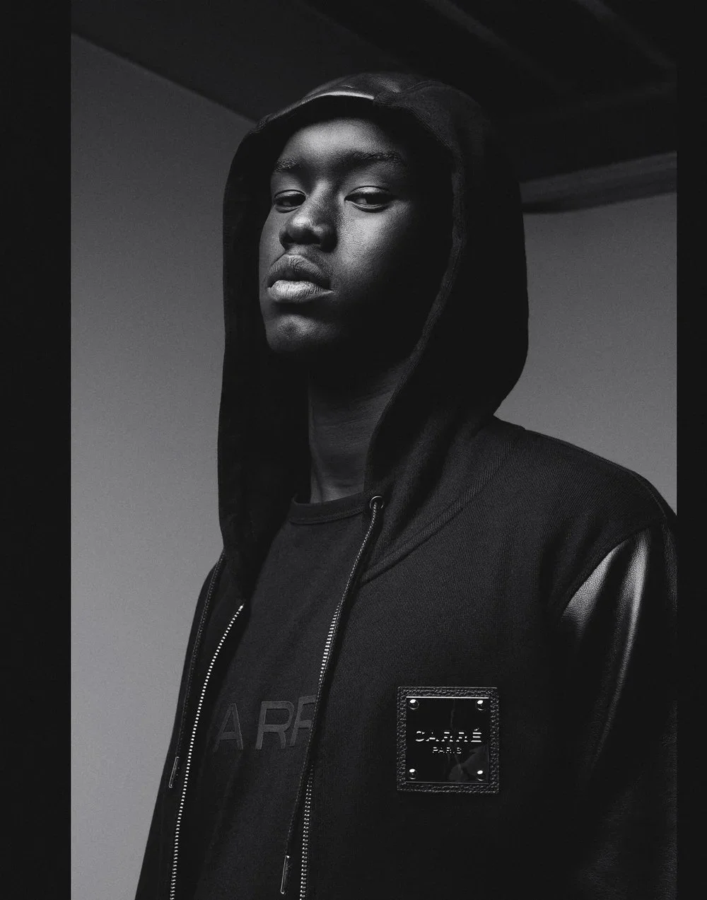 A young man with dark skin wearing a hooded black jacket with leather sleeves, standing against a plain background.