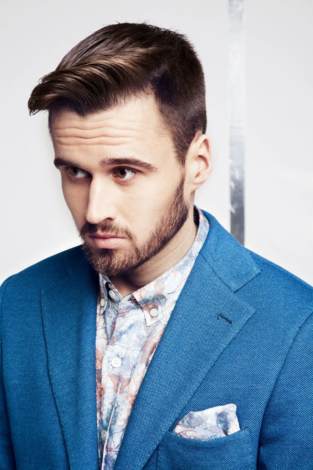 A man with dark brown hair styled with a side part and a beard, wearing a blue blazer over a patterned shirt, looking to the side with a serious expression.