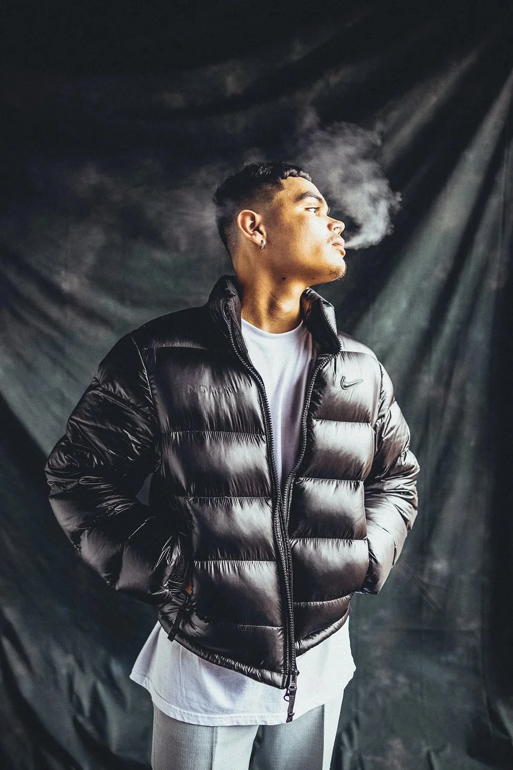 A young man with short hair and earrings wearing a black puffer jacket and white shirt, standing against a dark backdrop, exhaling smoke or vapor.