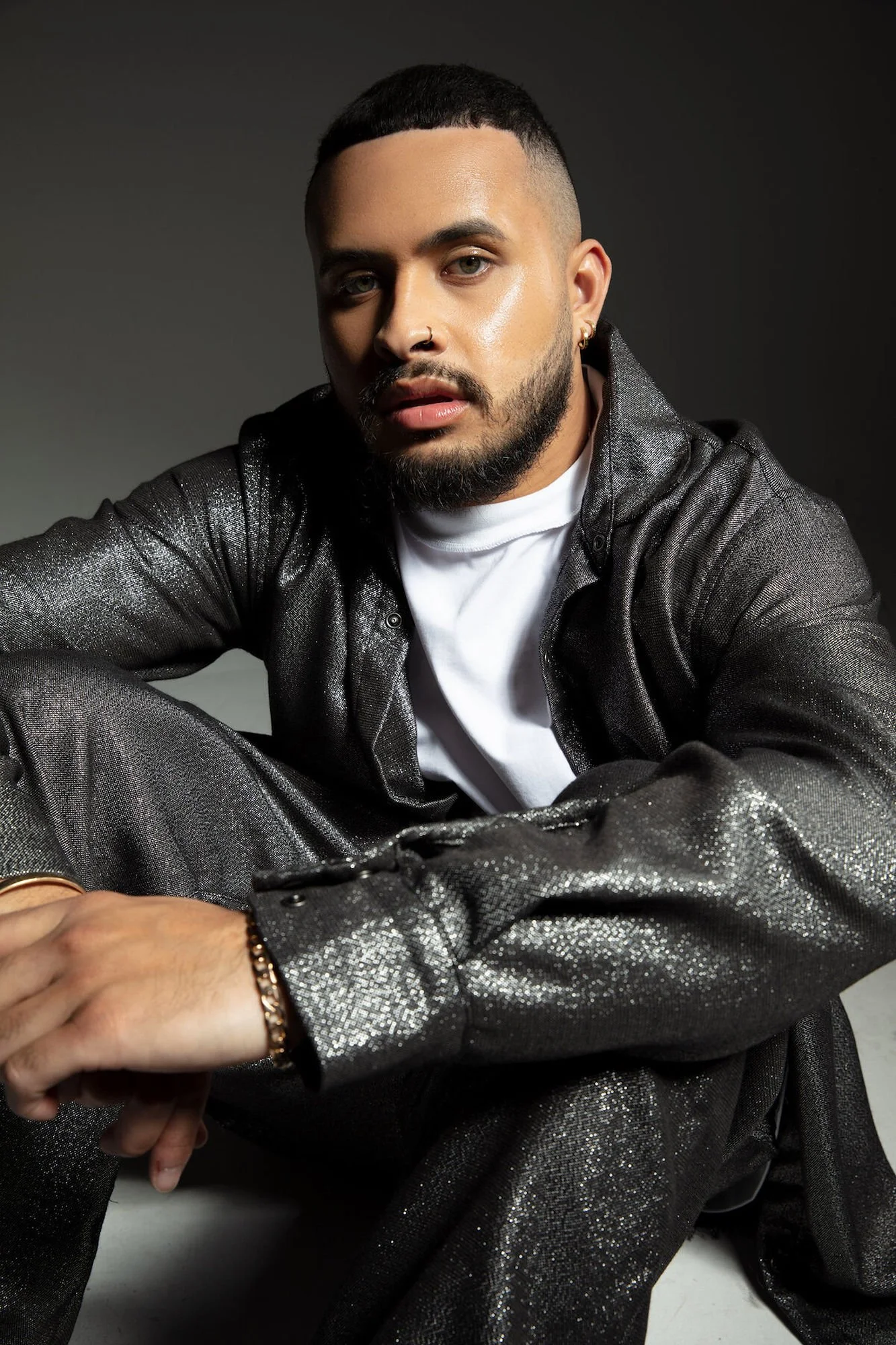 A man with a beard and short hair, wearing a shiny black suit and a white T-shirt, sitting in front of a dark background.