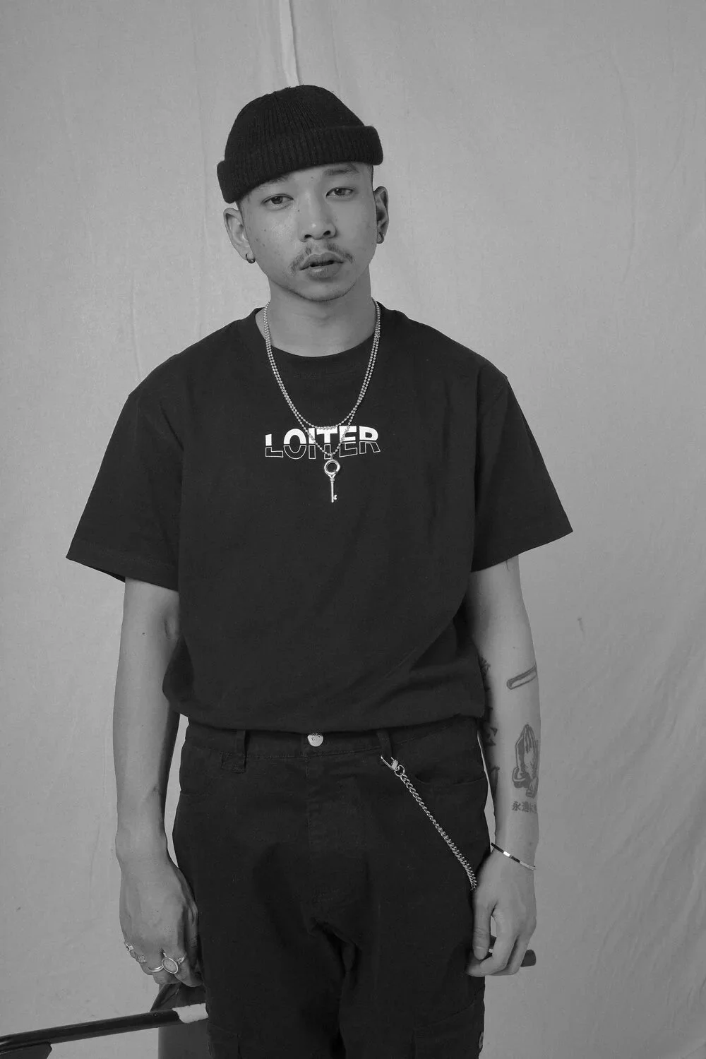 A young man wearing a black beanie, black T-shirt with the word 'LOITER' printed on it, chains, bracelet, rings, and tattoos, standing against a plain wall in black and white.