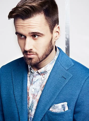 A young man with styled brown hair and a beard wearing a blue blazer over a floral shirt, looking to the side against a plain light background.
