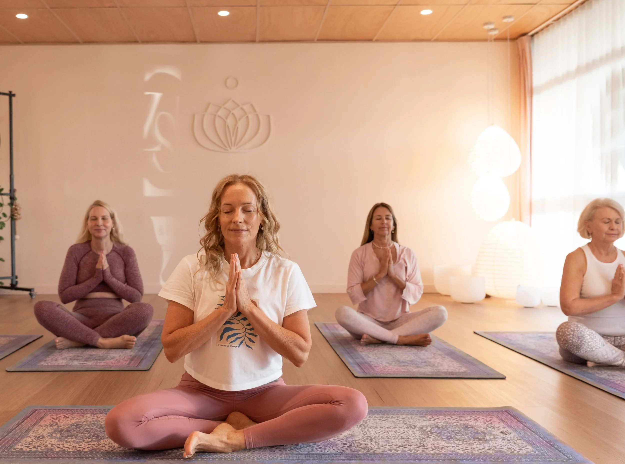 Four women practicing yoga or meditation in a serene indoor setting with warm lighting and mats on the wooden floor.