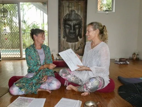 Two women sit cross-legged on a wooden floor, engaging in a conversation and smiling. One woman holds papers, and the background includes a large Buddha painting and windows showing greenery outside.