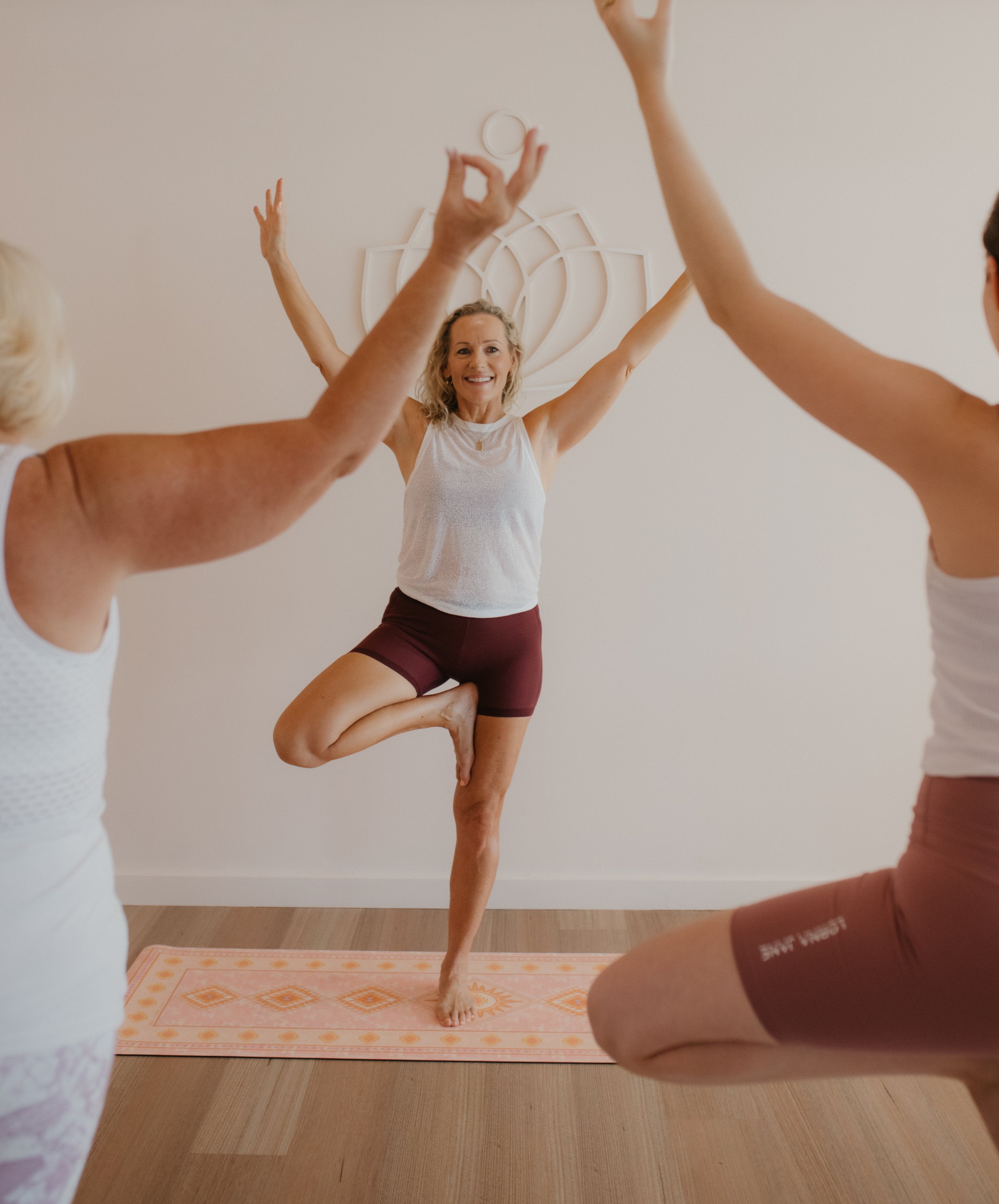 Women participating in a yoga class, with one woman balancing on one foot in a tree pose, smiling, while others raise their arms around her.