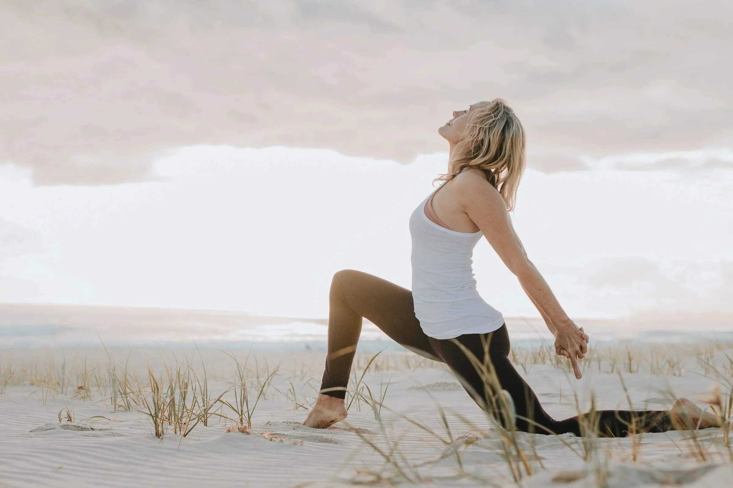 Woman practicing yoga on sandy beach at sunset, wearing a white tank top and black leggings, in a lunge pose with a high back arch, eyes closed, and arm extended behind her.